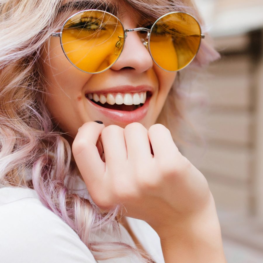 Close-up portrait of attractive joyful girl with amazing smile wears stylish yellow sunglasses and white shirt. Stunning curly young lady with blonde hair excitedly posing on the street.